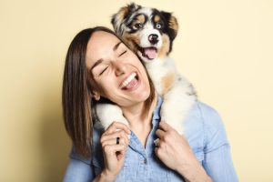 Siling woman with Australian Shepherd puppy looking over her shoulder on a yellow background. Showing one of the many benefits of dog ownership, happiness.